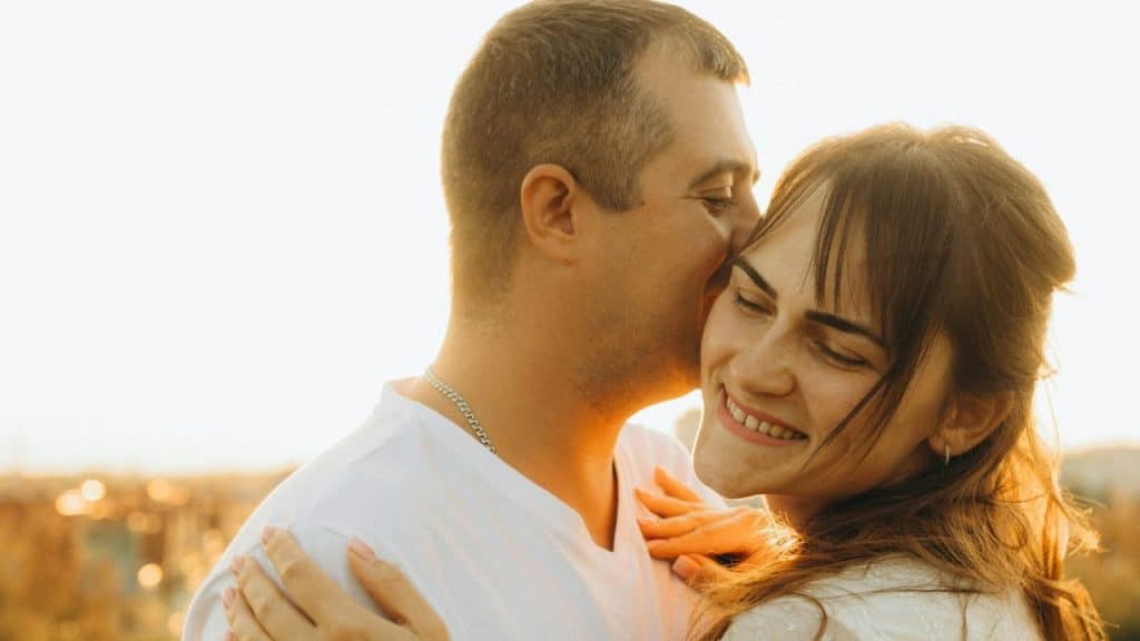 Man kissing a smiling woman on the cheek outdoors during sunset.