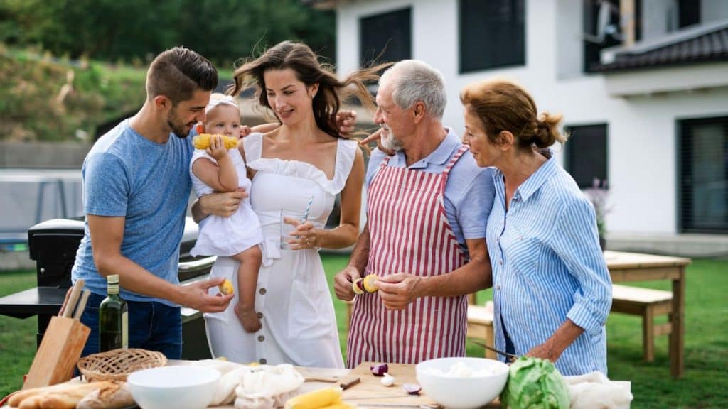 Three generations of a family having a barbecue in a yard in front of a house.