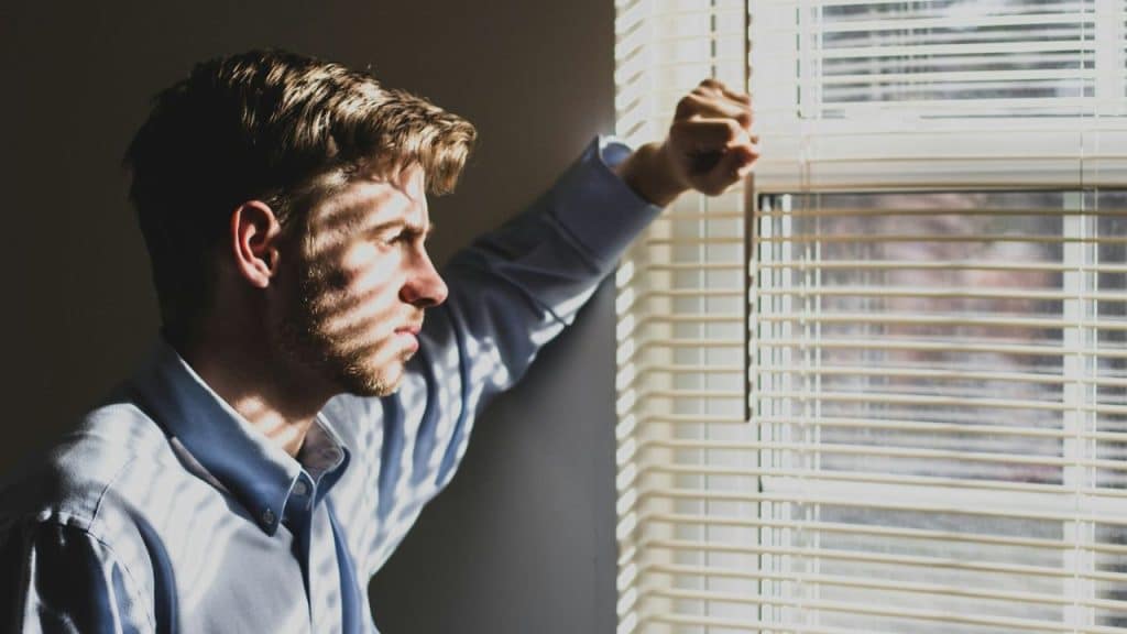 A man in a light blue shirt looks out a window with horizontal blinds, lit by dramatic sunlight.