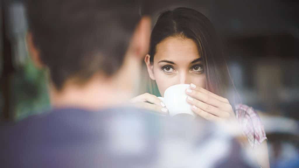 Woman looking up while drinking coffee across from a blurred man.
