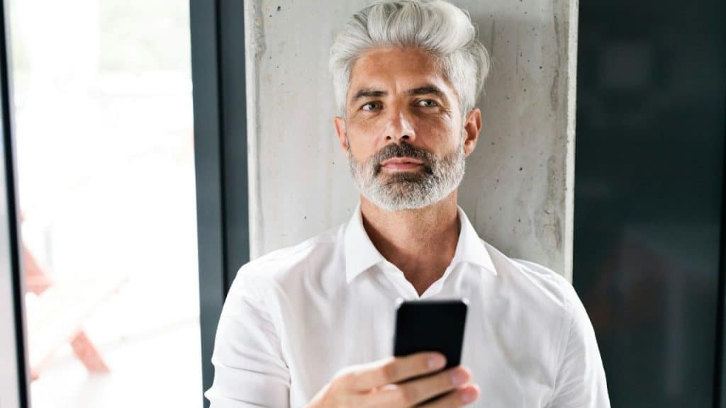 A mature man with gray hair and a beard holds a smartphone and looks at the camera.