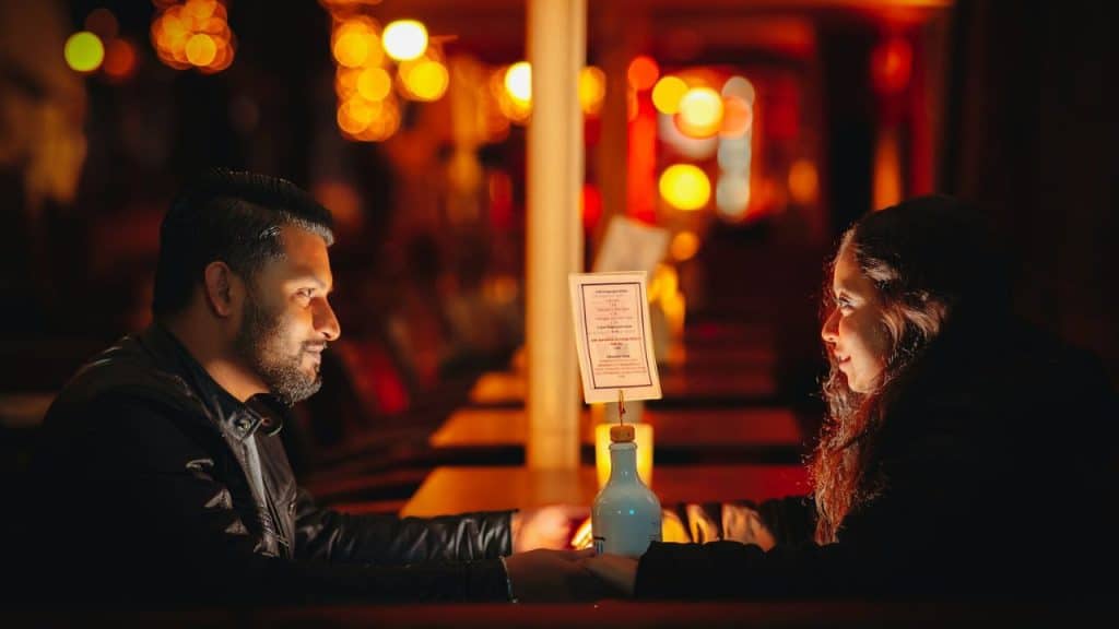 A smiling couple holds hands across a table in a dimly lit, warm-toned restaurant.