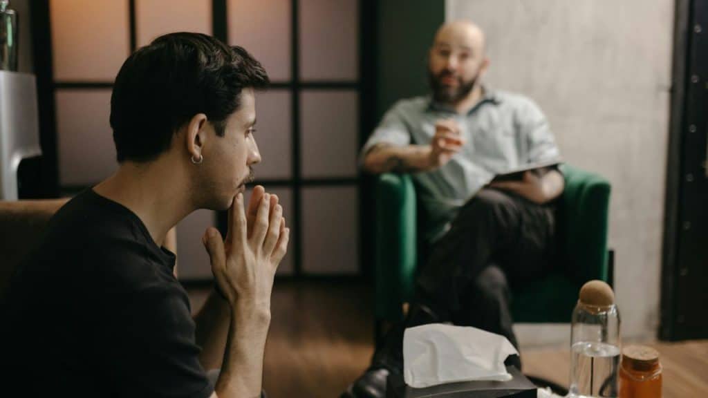 A man with hands clasped talks to a man sitting in an armchair.