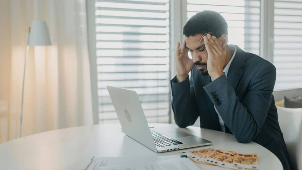 A man in a suit rubs his temples in frustration while looking at a laptop and documents.