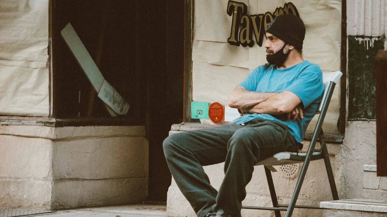 A man in a blue shirt, black beanie, and beard sits outdoors on a folding chair.