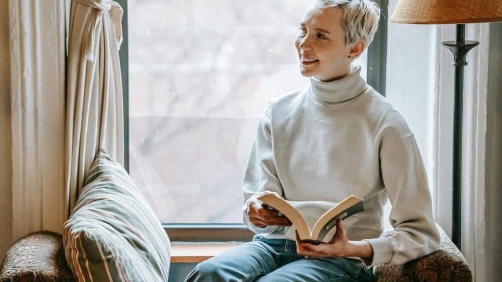 A smiling woman sits by a window holding an open book.