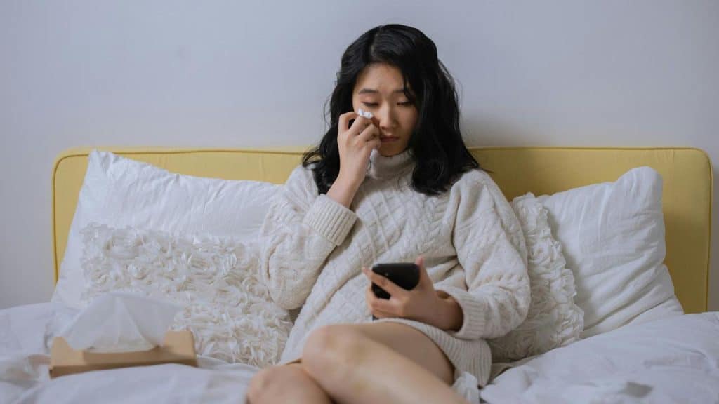 A woman sitting on a bed wiping her tears while looking at her phone.