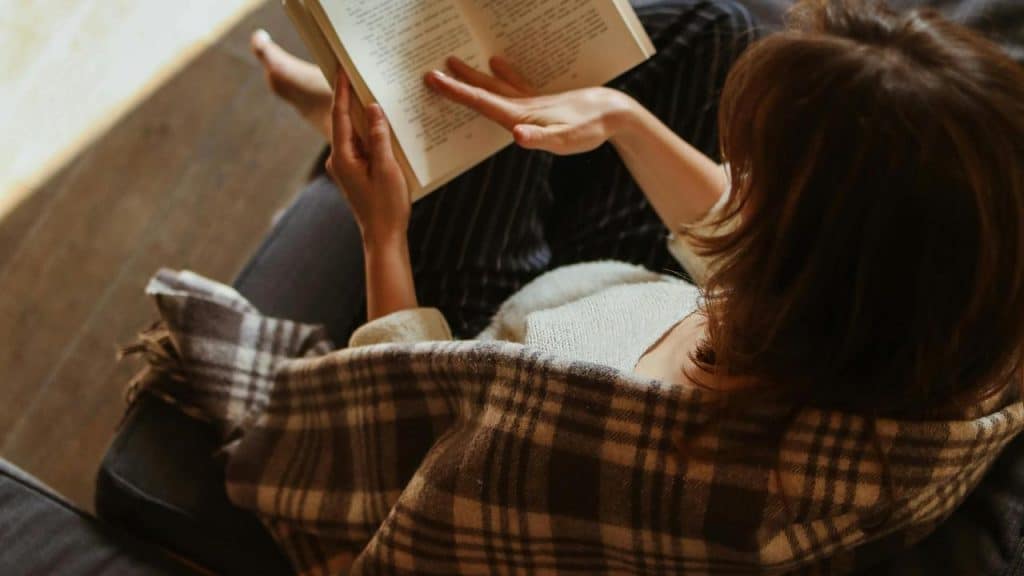 A woman reading a book while wrapped in a plaid blanket on a couch.