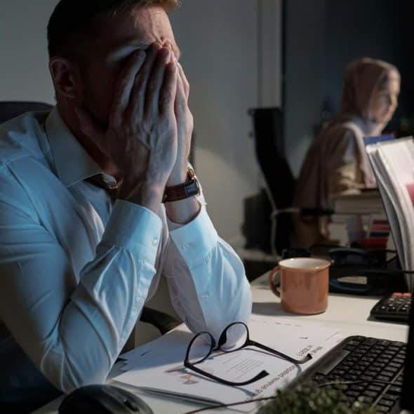 A stressed man in an office covering his face with his hands while working late.