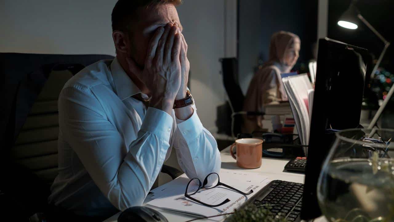 A stressed man in an office covering his face with his hands while working late.