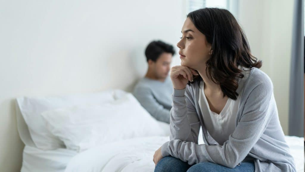 A woman looking sad while sitting on a bed as her partner sits on the bed in the background.