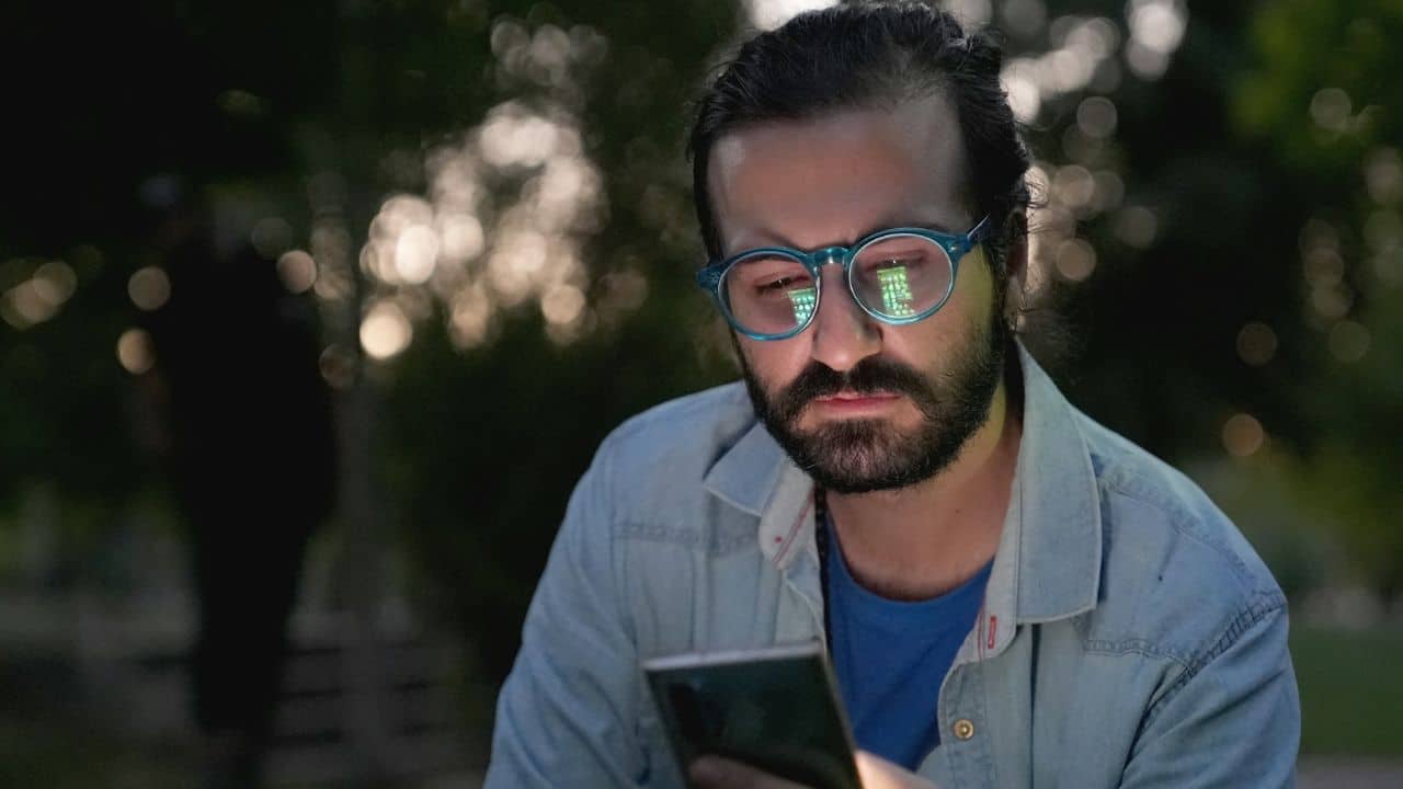 A man with a beard and glasses looking down at a phone outdoors at night.