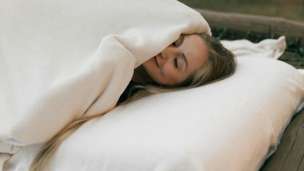 A woman smiling in bed while partly covered by a blanket.