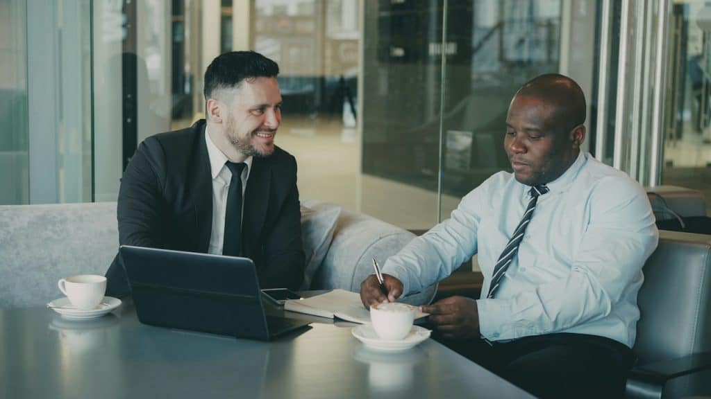 Two businessmen are sitting at a table in a modern setting, with one smiling at the other who is writing in a notebook.