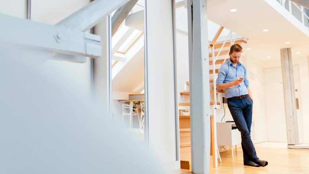A man in a blue shirt stands in a bright modern loft, looking at his phone.