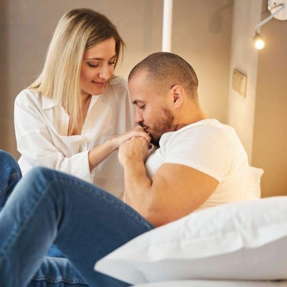 A man kisses a blonde woman's hand while sitting together on a bed.