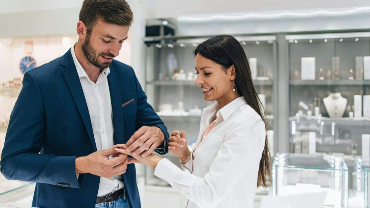 A man is placing a ring on a smiling woman's finger inside a jewelry store.
