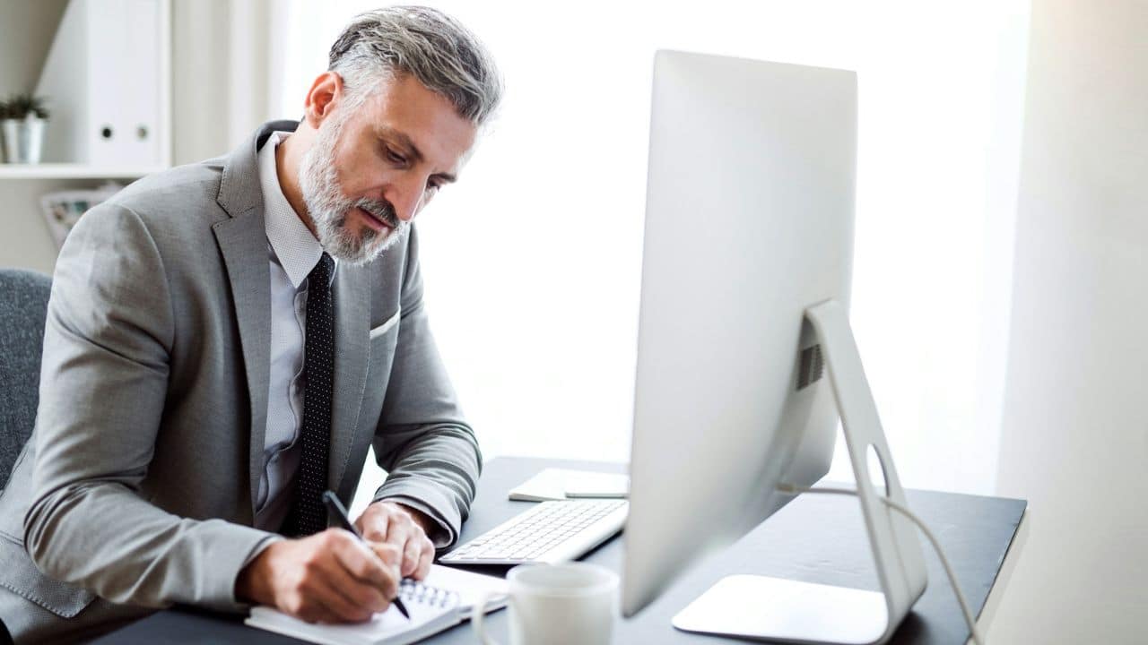 A well-dressed, gray-haired man writing in a notebook at a modern office desk.