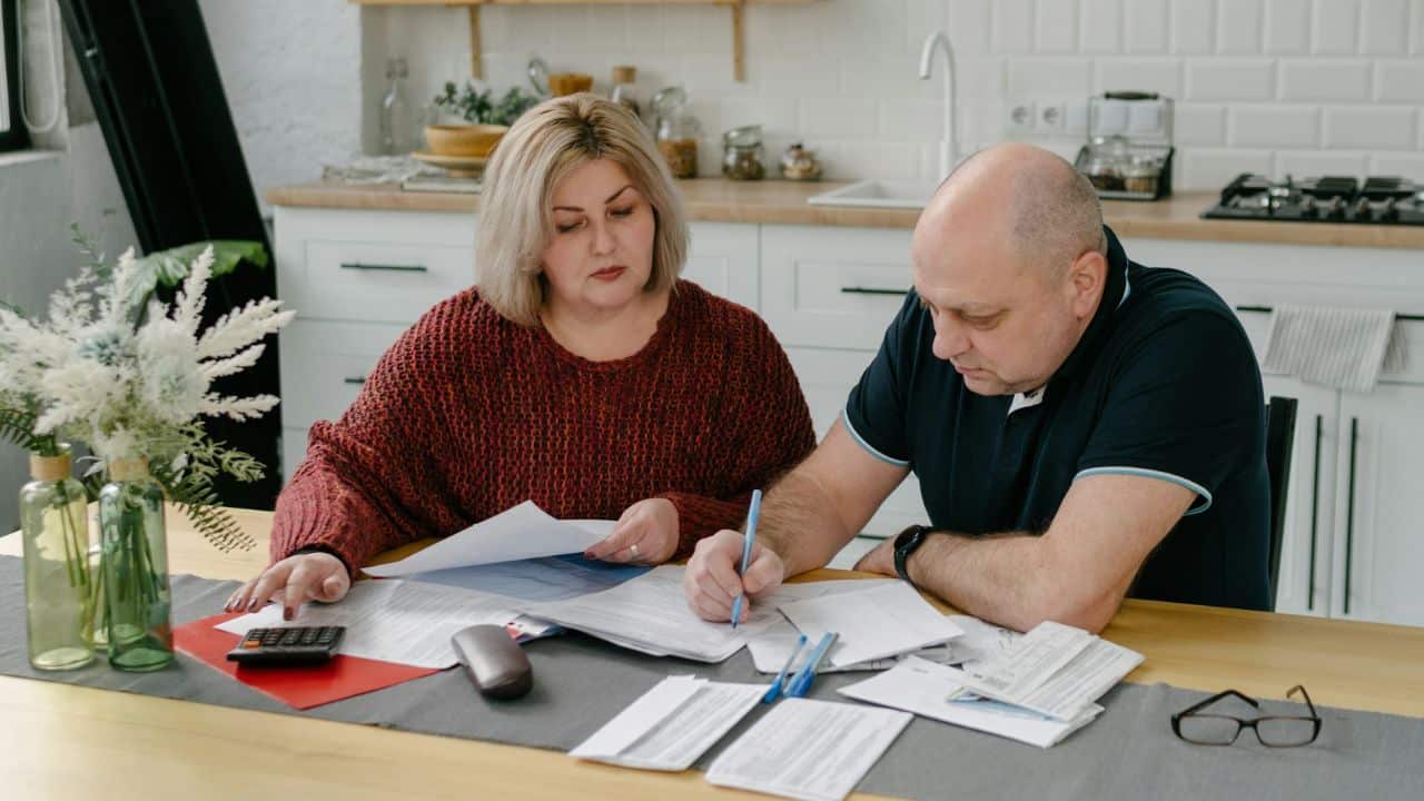 A middle-aged man and woman review papers and a calculator at a kitchen table.