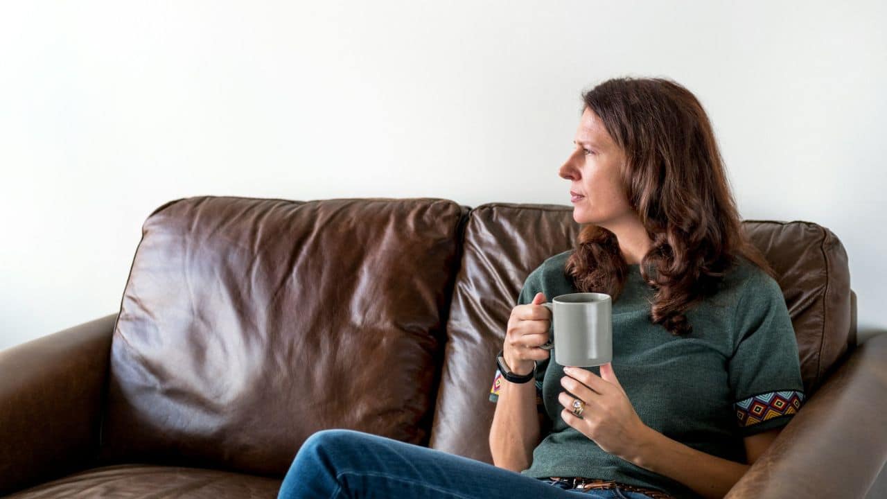 A woman sits on a brown leather couch holding a grey mug, looking away.