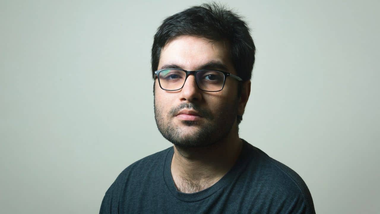 A man with a beard and glasses wears a dark shirt and looks forward.