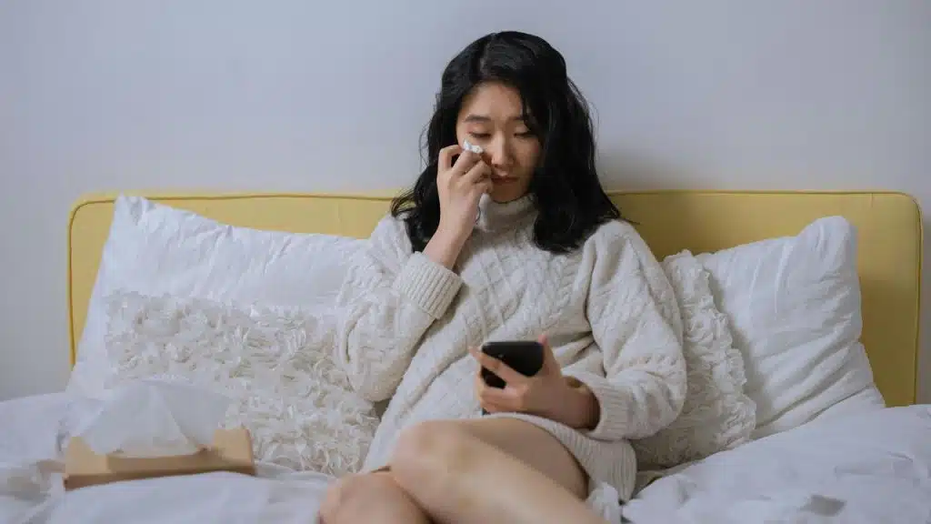 A woman sitting on a bed wiping her tears while looking at her phone.