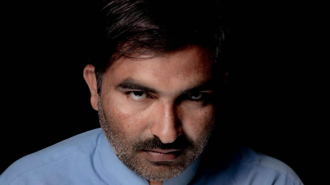 A man in a blue shirt with an intense stare is dimly lit against a black background.