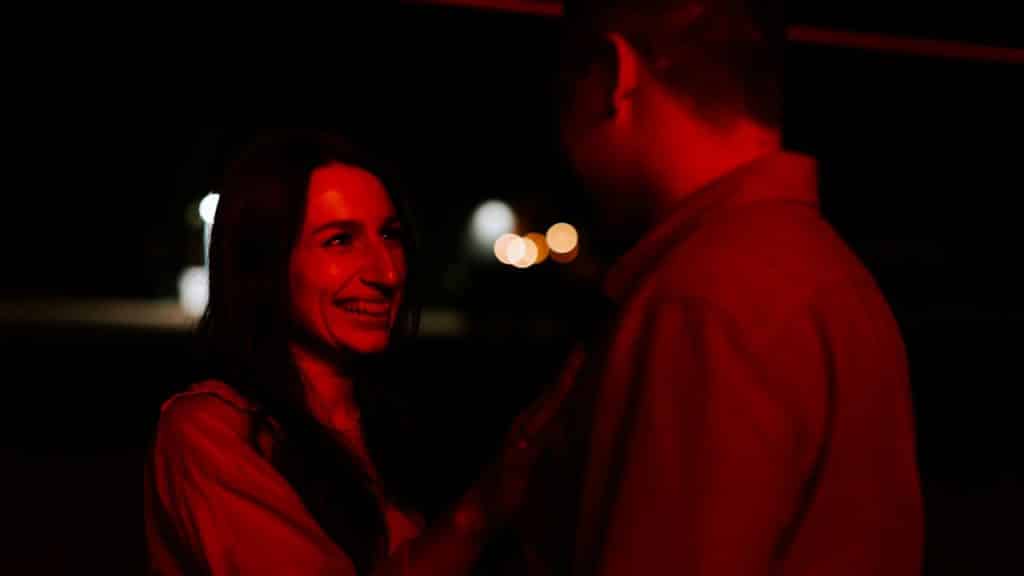 A smiling woman is looking up at a man in a dimly lit, red-hued setting.