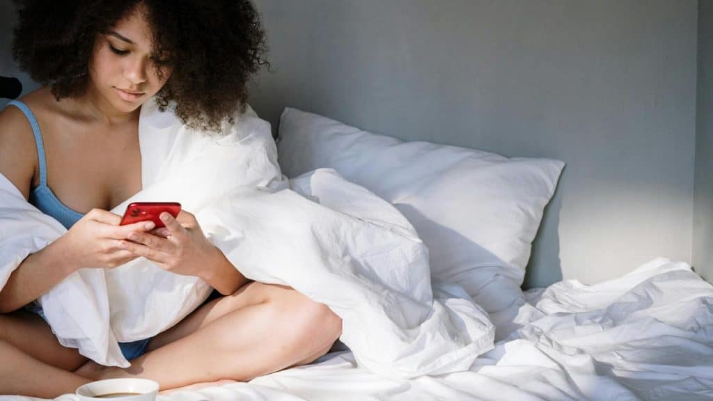 A woman sitting on a bed wrapped in a blanket while using her phone.
