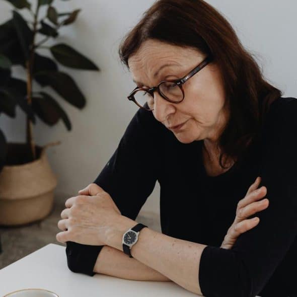 A woman with glasses sits at a table looking down thoughtfully.