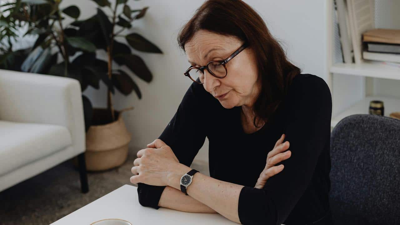 A woman with glasses sits at a table looking down thoughtfully.