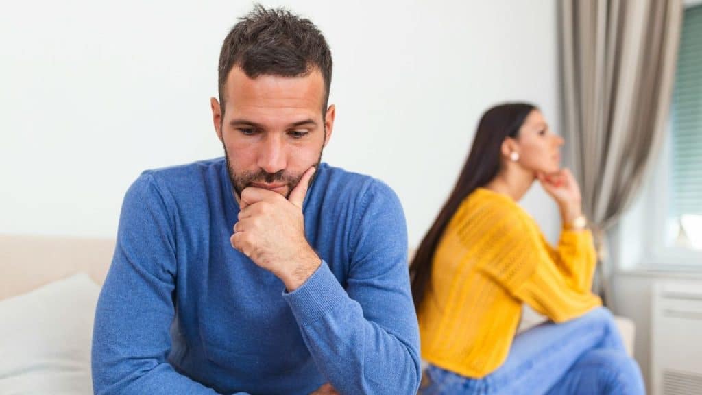 Man in blue sweater looking down, woman in yellow sweater blurred in the background.