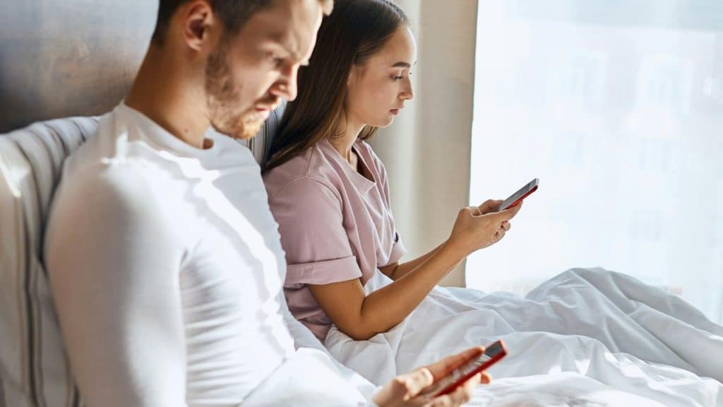 A man and a woman sit in bed, both looking down at their cell phones.