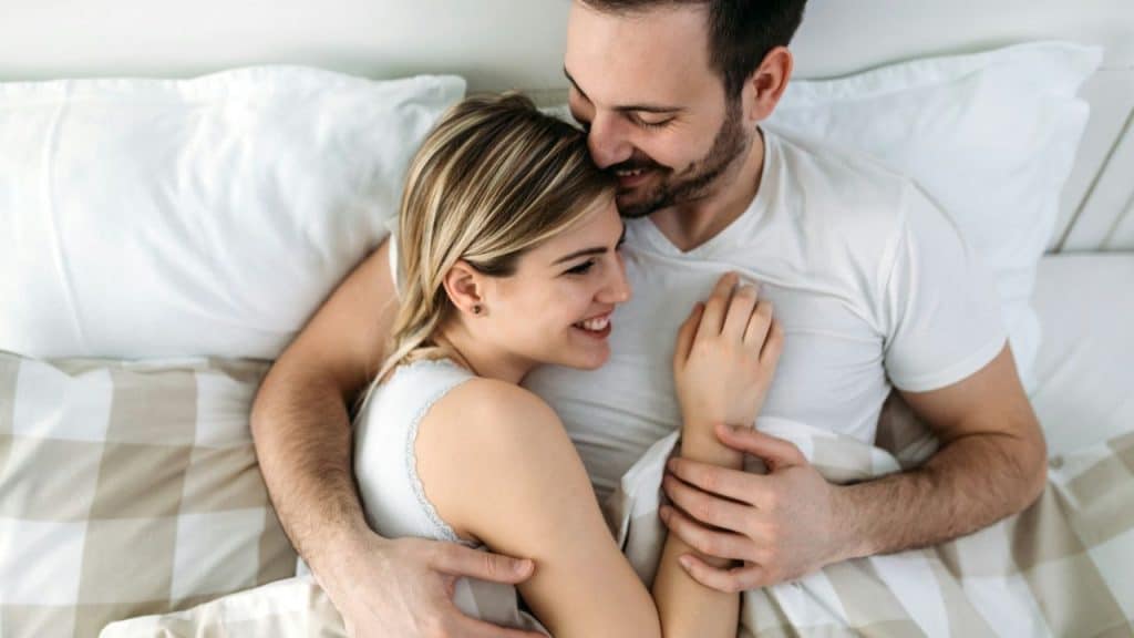 Smiling couple lying in bed embracing under a white and tan comforter.
