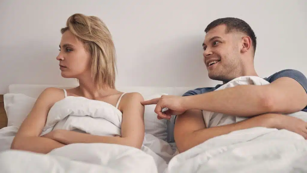 A smiling man pokes a finger at a woman who sits with her back turned in bed.
