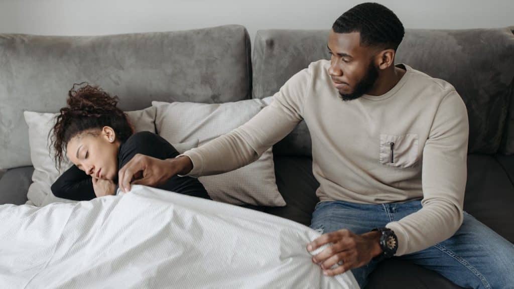 A man is sitting next to a woman who is asleep in bed, gently pulling a blanket over her.