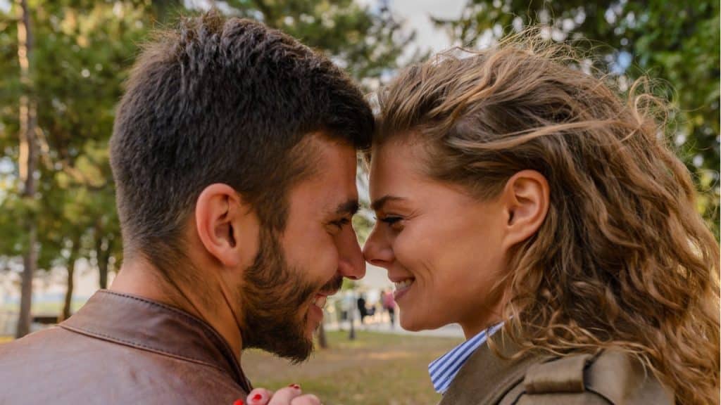 A smiling couple stands close together outdoors, touching foreheads and holding hands, viewed in a close-up profile shot.