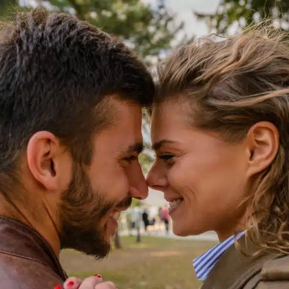 A smiling couple stands close together outdoors, touching foreheads and holding hands, viewed in a close-up profile shot.