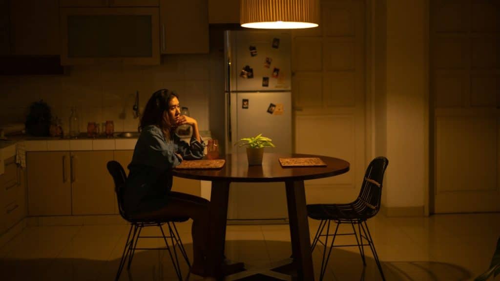 A woman is sitting alone at a kitchen table in a dimly lit room, resting her chin on her hands.