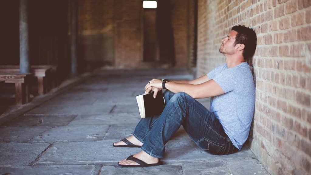 Man leaning against a brick wall, sitting on the ground holding a book.
