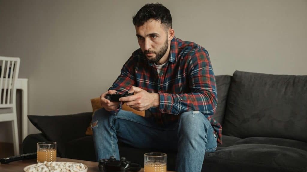 Man in a plaid shirt and jeans playing a video game on a gray couch.