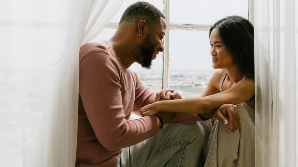 Bearded man and woman sitting by a window, holding hands and smiling at each other.