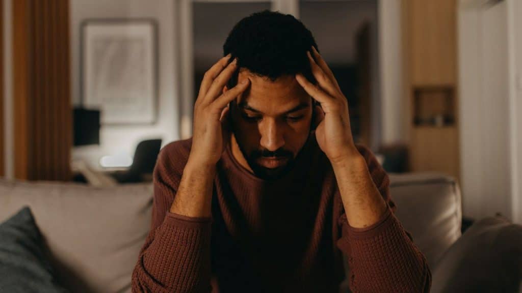 A stressed man sits in a dimly lit room, holding his head with both hands.