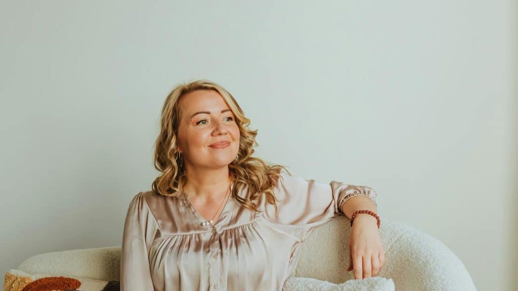 A woman sits comfortably on a couch, smiling softly and looking to the side.