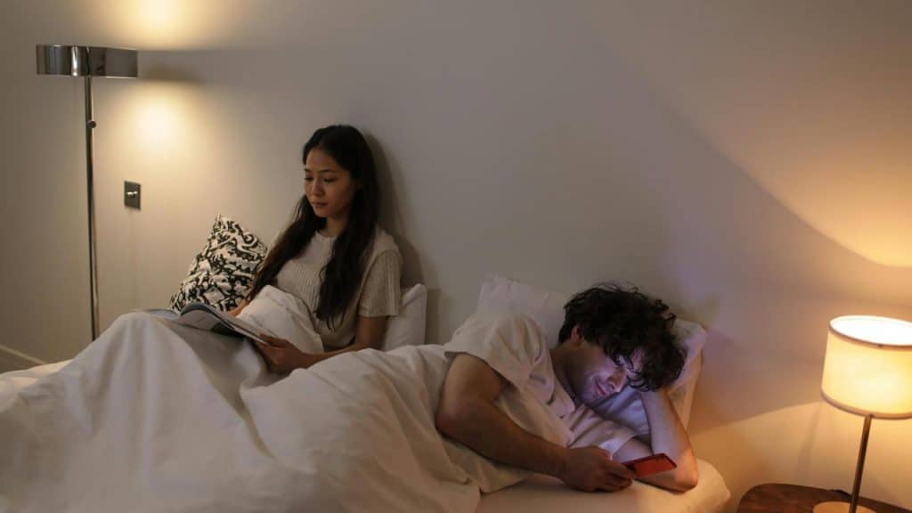 A couple lying in bed, one reading a book and the other using a phone.
