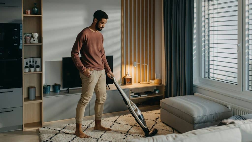 A casually dressed, barefoot man is vacuuming a patterned rug in a modern, sunlit living room.