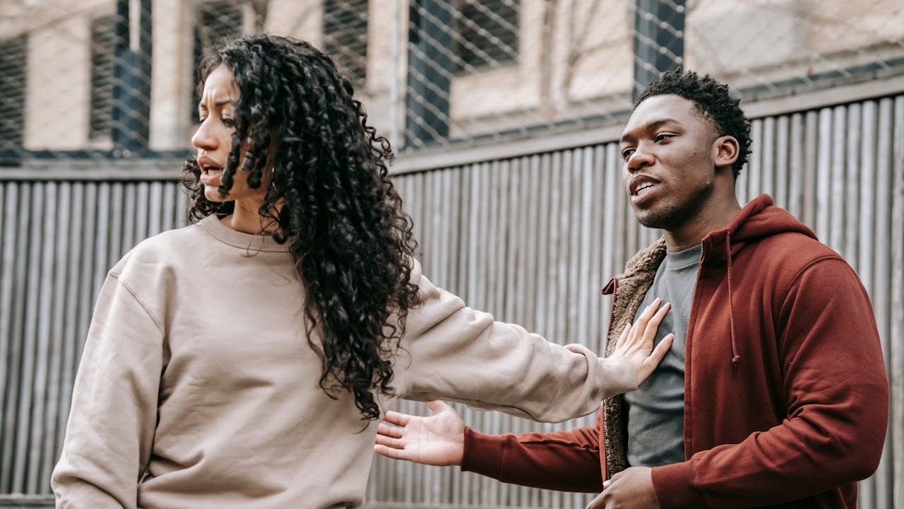 A young couple arguing outdoors, with the woman pushing the man away with her arm while she looks away.