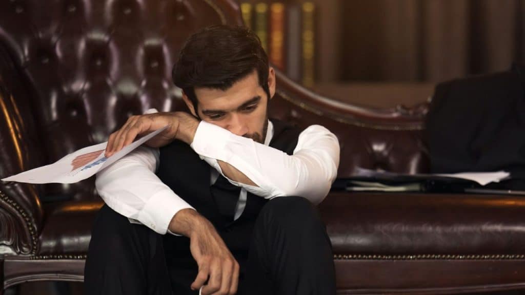A man in formal attire sits on the floor with his head down, leaning on a leather couch.