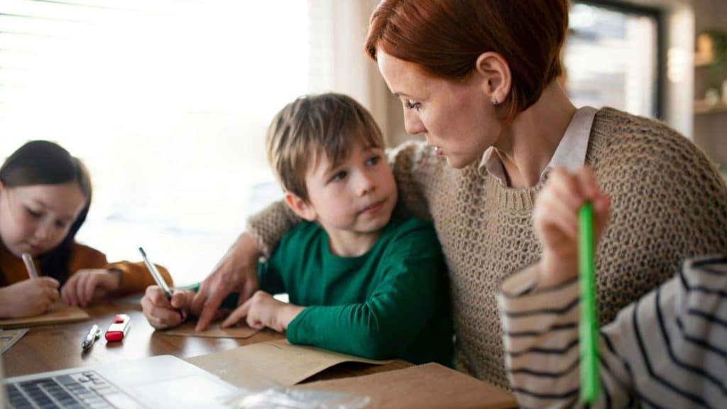Mother helping her son with schoolwork at a kitchen table while another child draws nearby.
