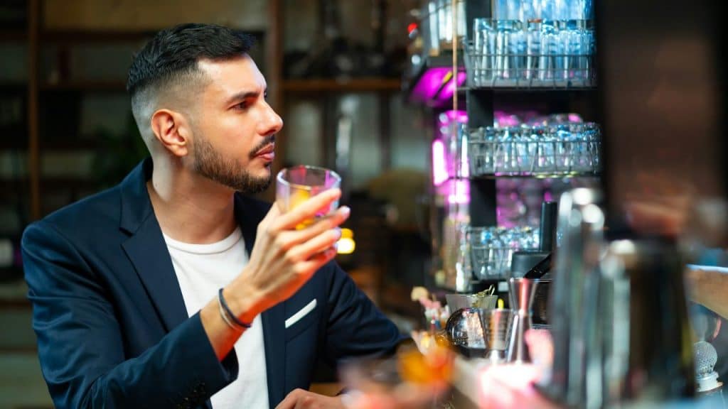 A well-dressed man holding a glass sits alone at a brightly lit bar counter.